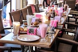 A table set with an array of milkshakes, each in a tall glass with a straw, surrounded by plates of pastries and colorful dot-patterned boxes. The table has a variety of condiments and glasses of water, and wicker chairs are arranged around it.