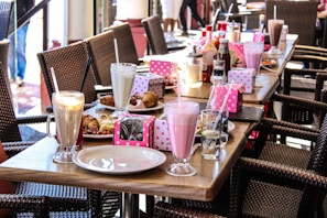 A table set with an array of milkshakes, each in a tall glass with a straw, surrounded by plates of pastries and colorful dot-patterned boxes. The table has a variety of condiments and glasses of water, and wicker chairs are arranged around it.