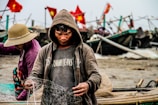 A person in a hoodie is focused on handling a fishing net, with fishing boats in the background decorated with red flags. Another person wearing a sun hat and purple jacket appears to be working alongside. The scene conveys a sense of hard work near a shore, likely engaged in fishing activities.