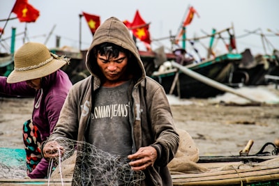 A person in a hoodie is focused on handling a fishing net, with fishing boats in the background decorated with red flags. Another person wearing a sun hat and purple jacket appears to be working alongside. The scene conveys a sense of hard work near a shore, likely engaged in fishing activities.