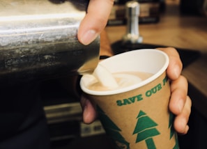 A person is pouring a creamy liquid, likely milk or cream, into a paper cup filled with coffee or hot chocolate. The cup has green illustrations of trees and text that promotes an environmental message. The setting appears to be a coffee shop or similar environment.