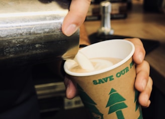 A person is pouring a creamy liquid, likely milk or cream, into a paper cup filled with coffee or hot chocolate. The cup has green illustrations of trees and text that promotes an environmental message. The setting appears to be a coffee shop or similar environment.