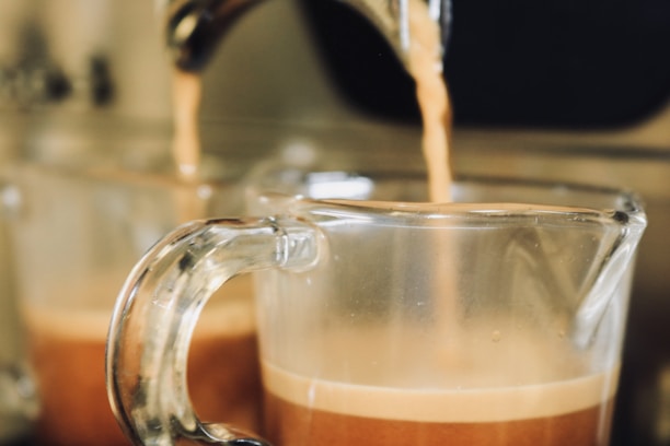 Two glass cups are being filled with espresso from a coffee machine. The liquid is a rich brown color with a layer of crema on top. The focus is on the front cup while the background is slightly blurred, emphasizing the process of coffee making.