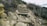 A rugged hiking trail with uneven wooden steps set into a rocky hillside. The path is surrounded by dry, sparse vegetation and bushes. Large rocks and shrubs line the sides of the trail, adding to the natural, untamed appearance of the landscape.