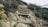A rugged hiking trail with uneven wooden steps set into a rocky hillside. The path is surrounded by dry, sparse vegetation and bushes. Large rocks and shrubs line the sides of the trail, adding to the natural, untamed appearance of the landscape.
