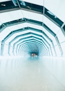 A modern architectural tunnel with an angular, geometric design. The structure is composed of repeating hexagonal patterns with a reflective surface giving a futuristic feel. The interior is brightly lit with a mix of natural and artificial light, creating a clean and orderly environment.