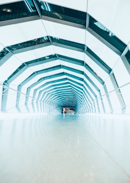 A modern architectural tunnel with an angular, geometric design. The structure is composed of repeating hexagonal patterns with a reflective surface giving a futuristic feel. The interior is brightly lit with a mix of natural and artificial light, creating a clean and orderly environment.