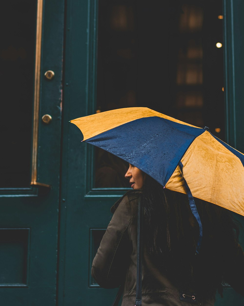 mulher sob o guarda-chuva amarelo e azul ao lado da porta de madeira verde