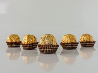 Five chocolate truffles wrapped in shiny gold foil are lined up on a reflective surface. Each piece is placed in a brown paper cup, and the lighting highlights the textures of the foil.