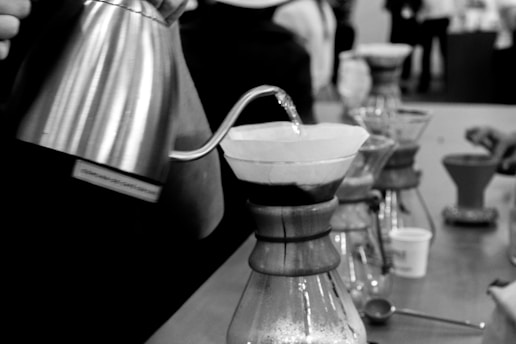 The image captures a close-up of a pour-over coffee brewing process, featuring a metallic kettle with a gooseneck spout pouring water into a dripper with a paper filter. Multiple chemex vessels are lined up on a countertop with various coffee-making paraphernalia visible in the background.