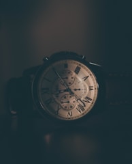 An elegant wristwatch with a classic design is displayed in dim lighting. The watch features a round face with Roman numerals and subdials. The texture and details of the watch are highlighted by the soft shadows and warm glow.