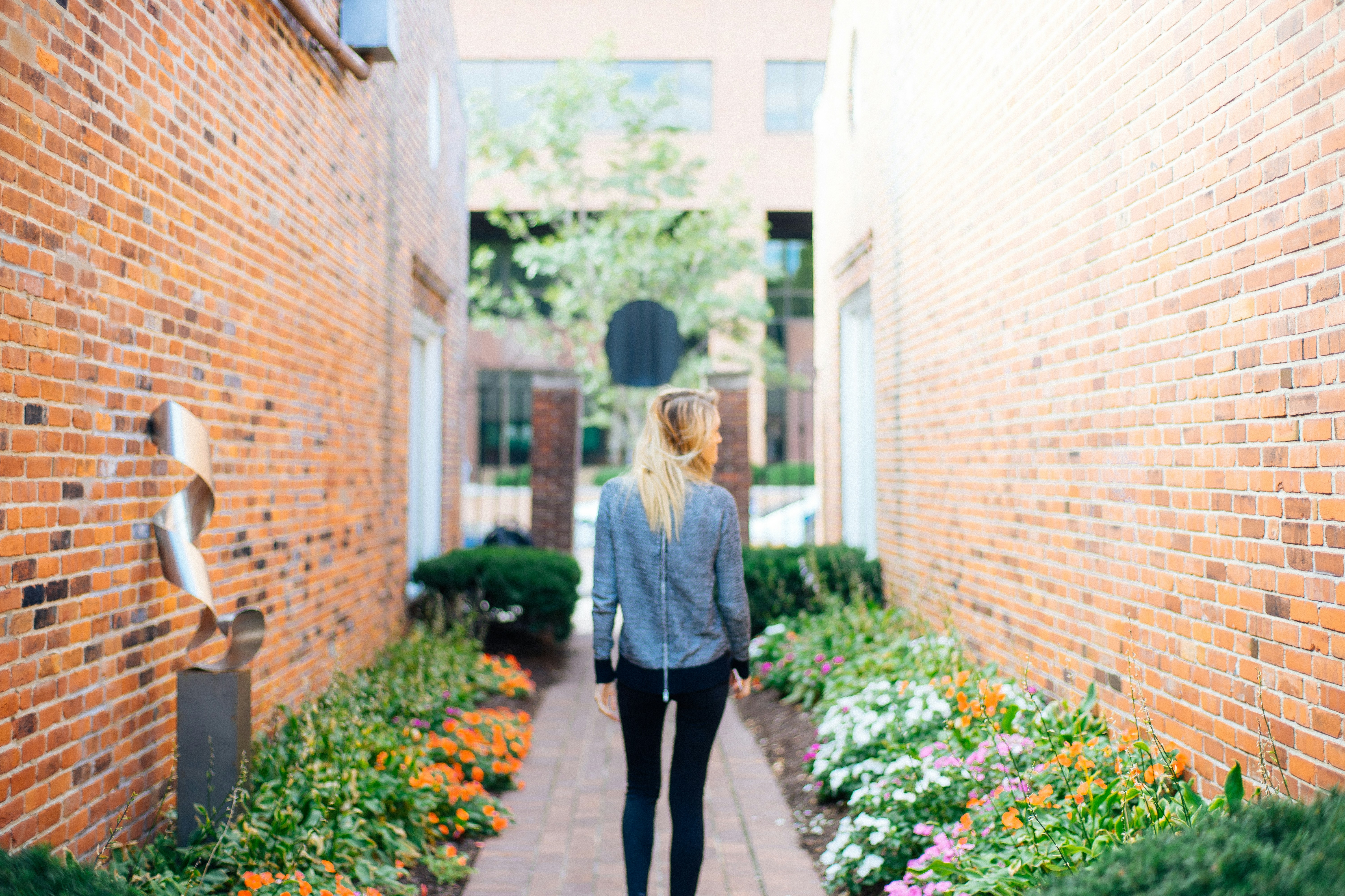 woman wearing gray long-sleeved shirt and black fitted pants in between of brown bricked walls with flowers during daytime