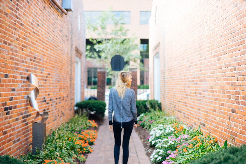 woman wearing gray long-sleeved shirt and black fitted pants in between of brown bricked walls with flowers during daytime