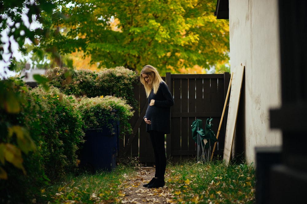 pregnant woman touching belly while looking at green bush near brown wooden fence during daytime