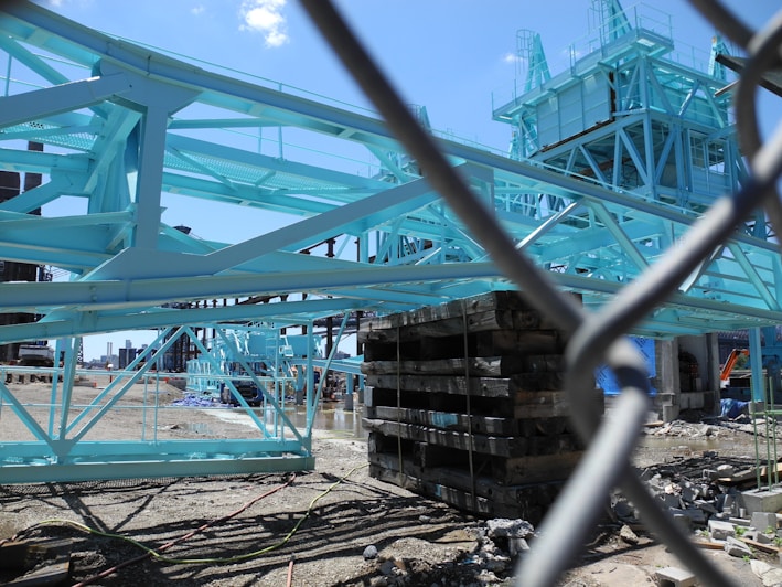 A construction site featuring a large structure with steel beams painted in light blue. Various building materials and equipment surround the site, and the area is fenced off with a chain-link fence. The sky is clear and blue, suggesting a sunny day.