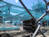 A construction site featuring a large structure with steel beams painted in light blue. Various building materials and equipment surround the site, and the area is fenced off with a chain-link fence. The sky is clear and blue, suggesting a sunny day.