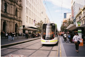 A modern tram travels along a city street surrounded by tall buildings and bustling pedestrians. The vehicle is predominantly white with some bright yellow and red markings. The street is lined with shops, and people are both walking and standing. Overhead wires crisscross above the tram, indicative of an urban transport network.