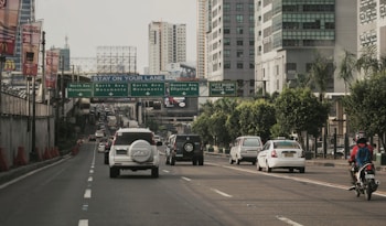 A busy urban road with multiple vehicles including cars and a motorcycle. High-rise buildings line both sides of the road. Above the road are multiple signboards providing directions. There are noticeable advertisements, including a McDonald's sign, on some of the buildings. The road is divided into several lanes with a noticeable flow of traffic.
