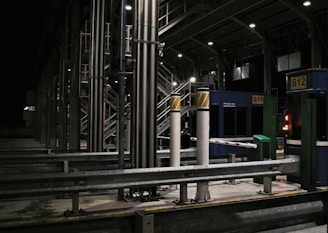 A toll booth area is depicted, featuring structures made of metal with numerous poles and booths marked with signs like S11 and S12. The setting is at night, illuminated by artificial lights, creating a stark contrast against the dark sky. The scene is largely empty with no visible vehicles or people.