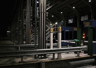A toll booth area is depicted, featuring structures made of metal with numerous poles and booths marked with signs like S11 and S12. The setting is at night, illuminated by artificial lights, creating a stark contrast against the dark sky. The scene is largely empty with no visible vehicles or people.