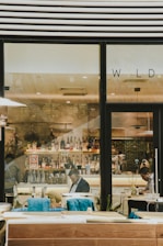 A modern bar setting with bottles of various drinks lined up on shelves behind the counter. A few people are seated, engaged in their activities, suggesting a relaxed environment. The decor includes wood and glass elements, creating a sleek and contemporary design.