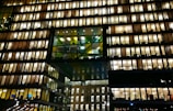 A modern office building with numerous floors, each floor lit up brightly with warm lighting. The windows reveal workspace interiors, with silhouettes of people and office furniture visible. The structure features a distinctive design with a central section of glass panels adding contrast.