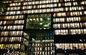 A modern office building with numerous floors, each floor lit up brightly with warm lighting. The windows reveal workspace interiors, with silhouettes of people and office furniture visible. The structure features a distinctive design with a central section of glass panels adding contrast.