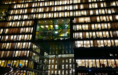A modern office building with numerous floors, each floor lit up brightly with warm lighting. The windows reveal workspace interiors, with silhouettes of people and office furniture visible. The structure features a distinctive design with a central section of glass panels adding contrast.