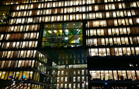 A modern office building with numerous floors, each floor lit up brightly with warm lighting. The windows reveal workspace interiors, with silhouettes of people and office furniture visible. The structure features a distinctive design with a central section of glass panels adding contrast.