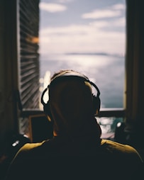 tilt-shift lens photography of person wearing headphones facing body of water through window of a dark room