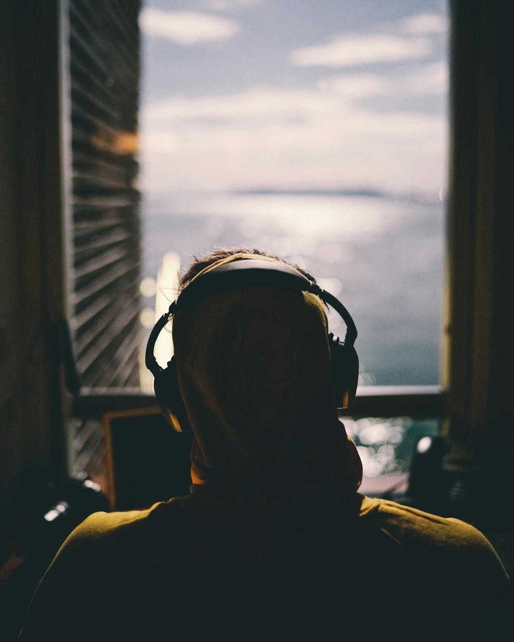 tilt-shift lens photography of person wearing headphones facing body of water through window of a dark room