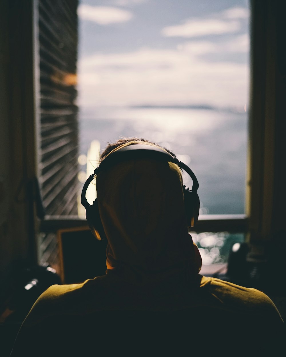 tilt-shift lens photography of person wearing headphones facing body of water through window of a dark room