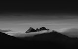 A dramatic black-and-white landscape featuring rugged mountain peaks rising above a blanket of clouds. The sky transitions from light to dark, creating a striking contrast with the silhouettes of the mountains. The image conveys a sense of vastness and solitude.