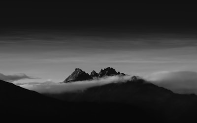 A dramatic black-and-white landscape featuring rugged mountain peaks rising above a blanket of clouds. The sky transitions from light to dark, creating a striking contrast with the silhouettes of the mountains. The image conveys a sense of vastness and solitude.
