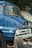 A vintage blue truck is parked next to some trees. The truck has a shiny chrome grille and round headlights, with a fabric draped over the front bumper. The background includes another vehicle partially covered by a tarp, and there are some fallen fruits on the ground.