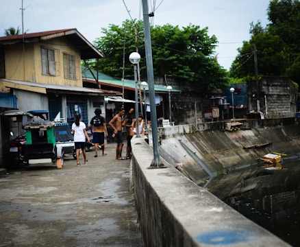 A group of people are gathered near a water canal or drain, next to a row of residential houses. The scene appears to be in a residential neighborhood with various structures visible. The people are casually dressed, some without shirts, suggestive of a warm climate. The area looks urban and somewhat rundown, with garbage bins and makeshift structures nearby.