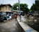 A group of people are gathered near a water canal or drain, next to a row of residential houses. The scene appears to be in a residential neighborhood with various structures visible. The people are casually dressed, some without shirts, suggestive of a warm climate. The area looks urban and somewhat rundown, with garbage bins and makeshift structures nearby.