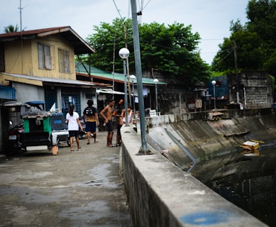 A group of people are gathered near a water canal or drain, next to a row of residential houses. The scene appears to be in a residential neighborhood with various structures visible. The people are casually dressed, some without shirts, suggestive of a warm climate. The area looks urban and somewhat rundown, with garbage bins and makeshift structures nearby.