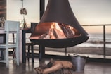 A modern, suspended fireplace with burning logs is set in a cozy indoor environment. Next to the fireplace, there is a wooden dining table with two chairs, one painted light blue. Various glass bottles and a couple of wine glasses are on the table. A bucket and some firewood sit on the floor near the fireplace. Large windows offer a view of the ocean and horizon during sunset, casting a warm glow over the room.