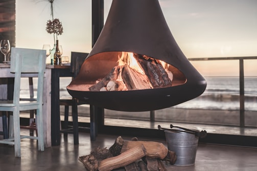 A modern, suspended fireplace with burning logs is set in a cozy indoor environment. Next to the fireplace, there is a wooden dining table with two chairs, one painted light blue. Various glass bottles and a couple of wine glasses are on the table. A bucket and some firewood sit on the floor near the fireplace. Large windows offer a view of the ocean and horizon during sunset, casting a warm glow over the room.