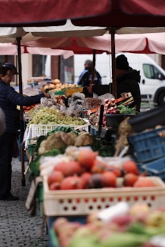 A bustling outdoor market featuring an array of colorful fruits and vegetables. Shoppers are browsing the fresh produce displayed in wooden crates under large umbrellas. The market stalls are filled with items like apples, grapes, and radishes, creating a vibrant and lively scene.