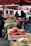 A bustling outdoor market featuring an array of colorful fruits and vegetables. Shoppers are browsing the fresh produce displayed in wooden crates under large umbrellas. The market stalls are filled with items like apples, grapes, and radishes, creating a vibrant and lively scene.