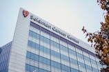 A modern building with reflective glass windows and a white exterior. The name 'Seidman Cancer Center' is prominently displayed at the top of the building. There is a logo to the left of the name, and the sky above is clear. Some tree branches with autumn-colored leaves are visible on the right side of the image.