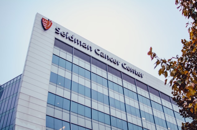 A modern building with reflective glass windows and a white exterior. The name 'Seidman Cancer Center' is prominently displayed at the top of the building. There is a logo to the left of the name, and the sky above is clear. Some tree branches with autumn-colored leaves are visible on the right side of the image.