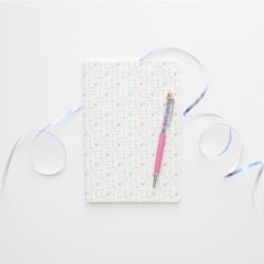 A notebook with a cover patterned with the word 'LOVE' repeatedly. It is accompanied by a pink pen laid across its surface. Light blue ribbons curl artistically around the notebook, all set against a clean, white background.