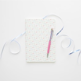 A notebook with a cover patterned with the word 'LOVE' repeatedly. It is accompanied by a pink pen laid across its surface. Light blue ribbons curl artistically around the notebook, all set against a clean, white background.