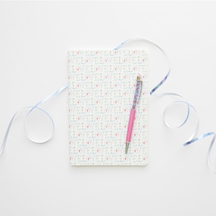 A notebook with a cover patterned with the word 'LOVE' repeatedly. It is accompanied by a pink pen laid across its surface. Light blue ribbons curl artistically around the notebook, all set against a clean, white background.