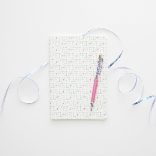 A notebook with a cover patterned with the word 'LOVE' repeatedly. It is accompanied by a pink pen laid across its surface. Light blue ribbons curl artistically around the notebook, all set against a clean, white background.