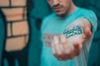 A person is extending their arm forward, displaying a tattoo on the forearm. The tattoo appears to depict intricate black designs, possibly featuring some animal or abstract shapes. The background is blurred, but it suggests an urban or graffiti setting with bold colors. The person's face is partially visible, but out of focus, emphasizing the tattoo and arm.