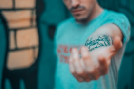 A person is extending their arm forward, displaying a tattoo on the forearm. The tattoo appears to depict intricate black designs, possibly featuring some animal or abstract shapes. The background is blurred, but it suggests an urban or graffiti setting with bold colors. The person's face is partially visible, but out of focus, emphasizing the tattoo and arm.